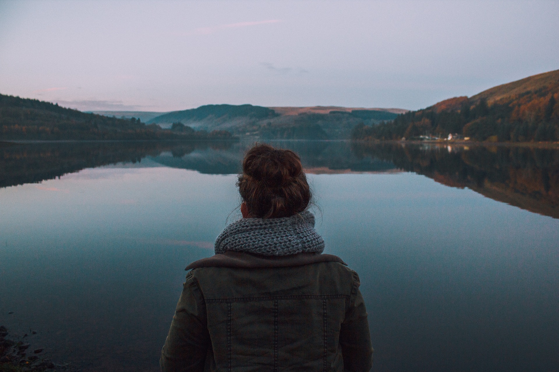 Une femme vêtu d'un manteau et d'un foulard regarde un lac et les montagnes