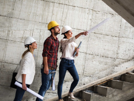 Trois personnes debout dans des marches avec un casque de construction suivant du regard une des 3 qui pointe au loin