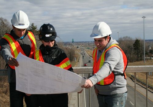 Trois personnes avec des chapeaux de sécurité sur une passerelle au-dessus d'une autoroute